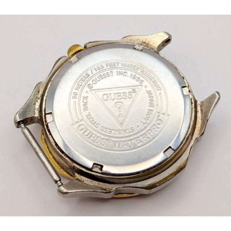Op de afbeelding: De achterkant van een zilveren en gouden Guess-horloge met een zwarte wijzerplaat. Het horloge is waterdicht tot 50 meter of 165 voet. Het horloge is gemaakt van roestvrij staal en is gemaakt in Japan.