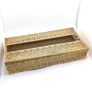 May include: A rectangular, gold-colored tissue box with intricate floral and scrollwork patterns. The box has a rectangular opening on top and a clear, brown-tinted interior. The design is ornate and vintage-inspired.