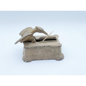 May include: A vintage, antique toothpick holder. The holder is a rectangular box with a bird-shaped lid. The bird is perched on top of the box, with its beak extended towards a toothpick. The holder is made of metal and has a weathered, aged appearance.