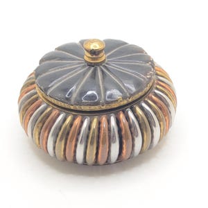 May include: A small, decorative trinket box with a lid. The lid is black with a gold knob, and the body is a mix of copper, silver, and gold-toned metal. The box has a ribbed design.