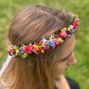 May include: A floral crown made of small, colorful flowers. The crown is worn on a person's head, with their brown hair visible.
