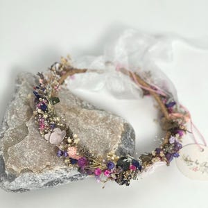 May include: A delicate floral crown made with dried flowers in shades of pink, purple, and white. The crown is made on a wire base and is adorned with small, delicate flowers and greenery.
