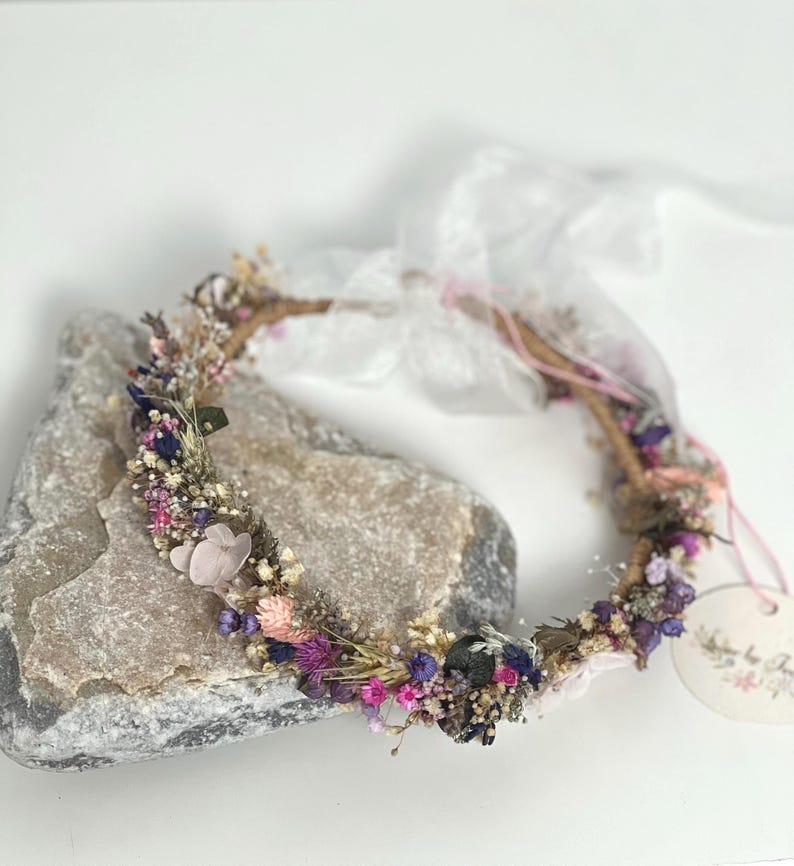 May include: A delicate floral crown made with dried flowers in shades of pink, purple, and white. The crown is made on a wire base and is adorned with small, delicate flowers and greenery.
