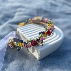 May include: A flower crown made with dried flowers in shades of pink, yellow, purple, and red. The crown is on a white surface.