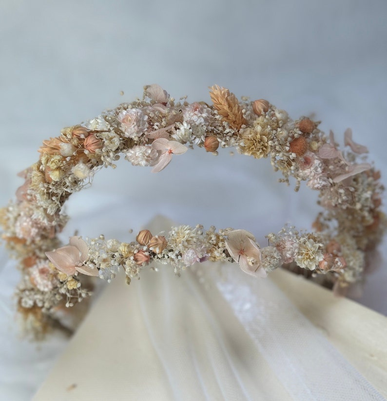Pode incluir: Uma coroa de flores secas delicada com forma circular. A coroa &eacute; feita de pequenas flores brancas e bege, com algumas flores maiores de cor p&ecirc;ssego espalhadas. A coroa &eacute; perfeita para um casamento ou outra ocasi&atilde;o especial.