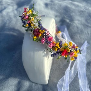 May include: A floral crown made with dried flowers in shades of pink, yellow, purple, and red. The crown is on a white surface and has a white ribbon attached.