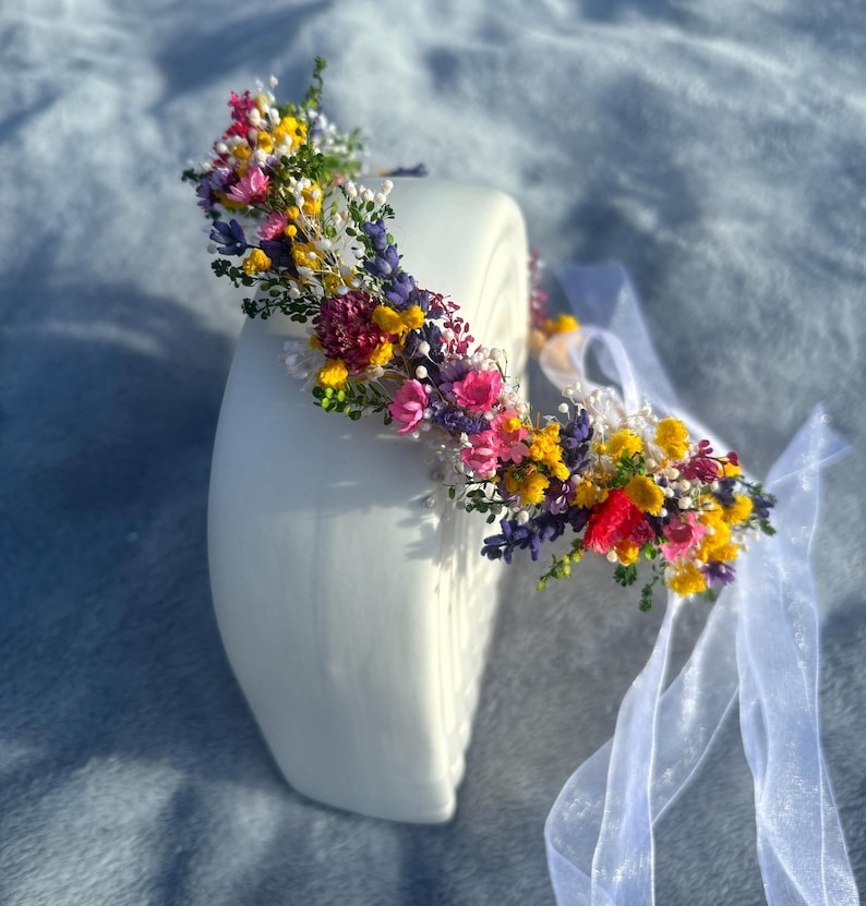 May include: A floral crown made with dried flowers in shades of pink, yellow, purple, and red. The crown is on a white surface and has a white ribbon attached.