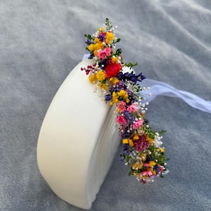 May include: A colorful flower crown with yellow, pink, purple, and white flowers. The crown is made of dried flowers and is sitting on a white surface.