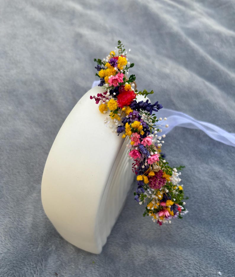 May include: A colorful flower crown with yellow, pink, purple, and white flowers. The crown is made of dried flowers and is sitting on a white surface.