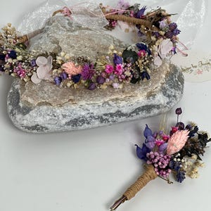 May include: A purple and pink dried flower crown and matching boutonniere. The crown is made with a variety of dried flowers, including lavender, pink roses, and baby's breath. The boutonniere is a smaller version of the crown, with a single pink rose and lavender.