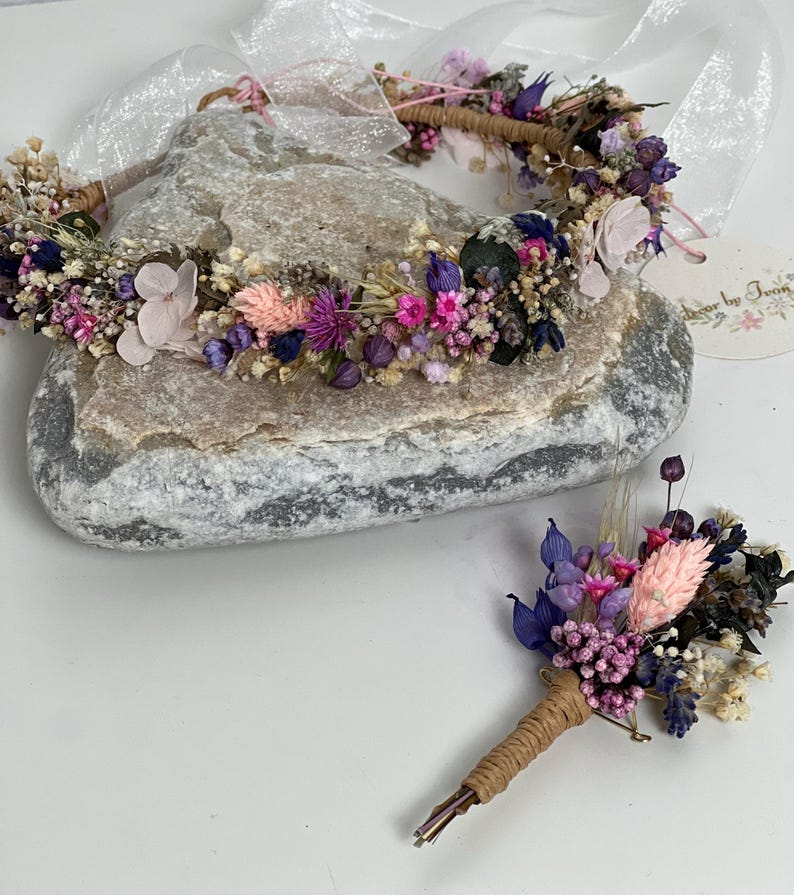 May include: A purple and pink dried flower crown and matching boutonniere. The crown is made with a variety of dried flowers, including lavender, pink roses, and baby's breath. The boutonniere is a smaller version of the crown, with a single pink rose and lavender.