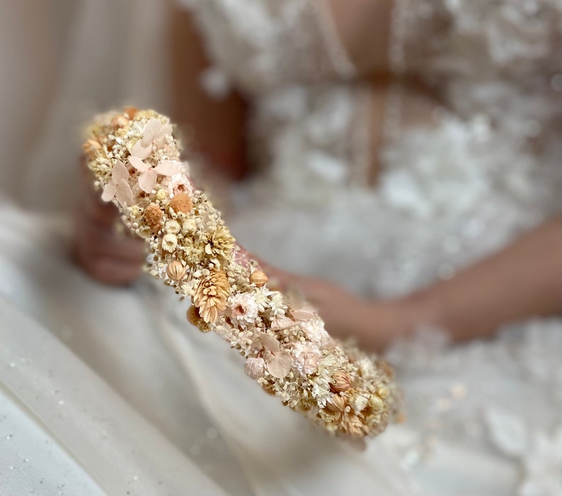 Pode incluir: Uma coroa floral delicada feita de flores secas em tons de creme, bege e rosa claro. A coroa &eacute; disposta em forma circular e apresenta uma variedade de pequenas flores e folhas.