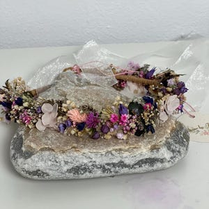 May include: A flower crown made with dried flowers in shades of pink, purple, and white. The crown is on a gray rock with a white tag that says "Flower by F".