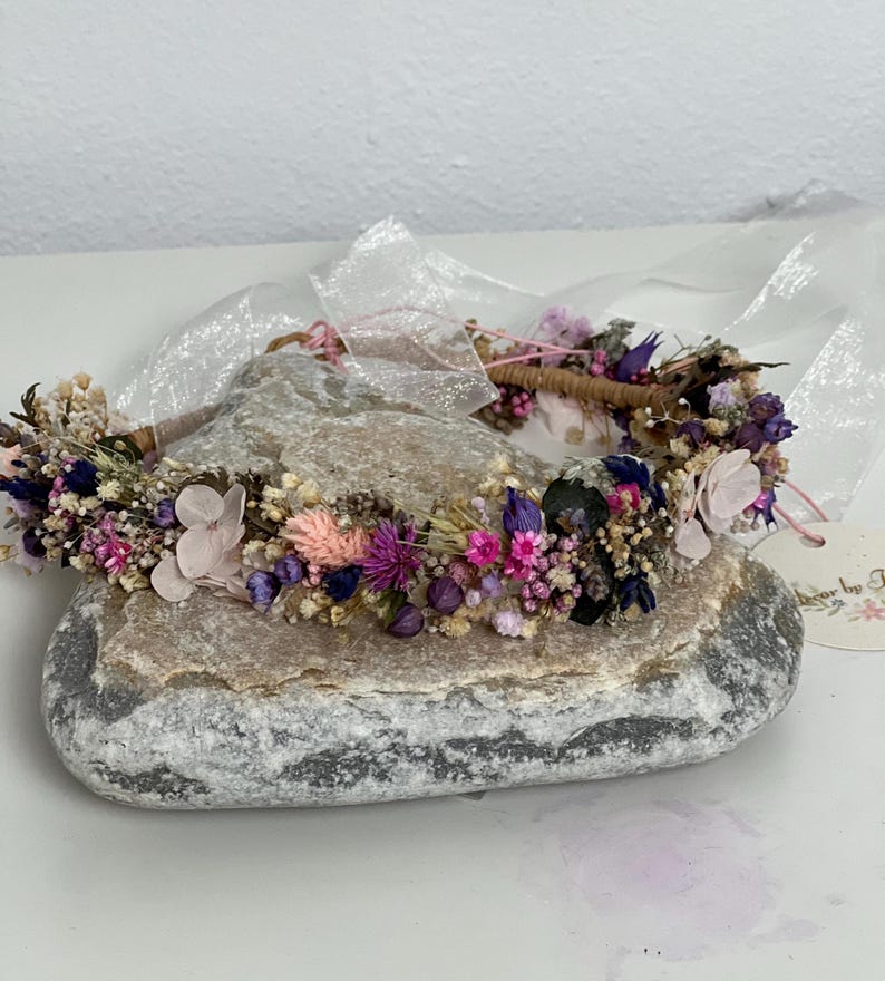 May include: A flower crown made with dried flowers in shades of pink, purple, and white. The crown is on a gray rock with a white tag that says "Flower by F".