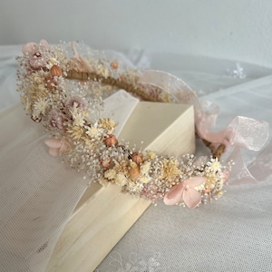 Pode incluir: Uma coroa floral delicada feita com flores secas em tons de rosa e bege. A coroa &eacute; adornada com pequenas flores rosa e gram&iacute;neas secas bege. &Eacute; amarrada com uma fita rosa.
