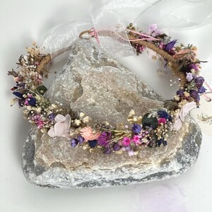 May include: A delicate floral crown made with dried flowers in shades of pink, purple, and white. The crown is wrapped in brown twine and has a light pink ribbon attached.