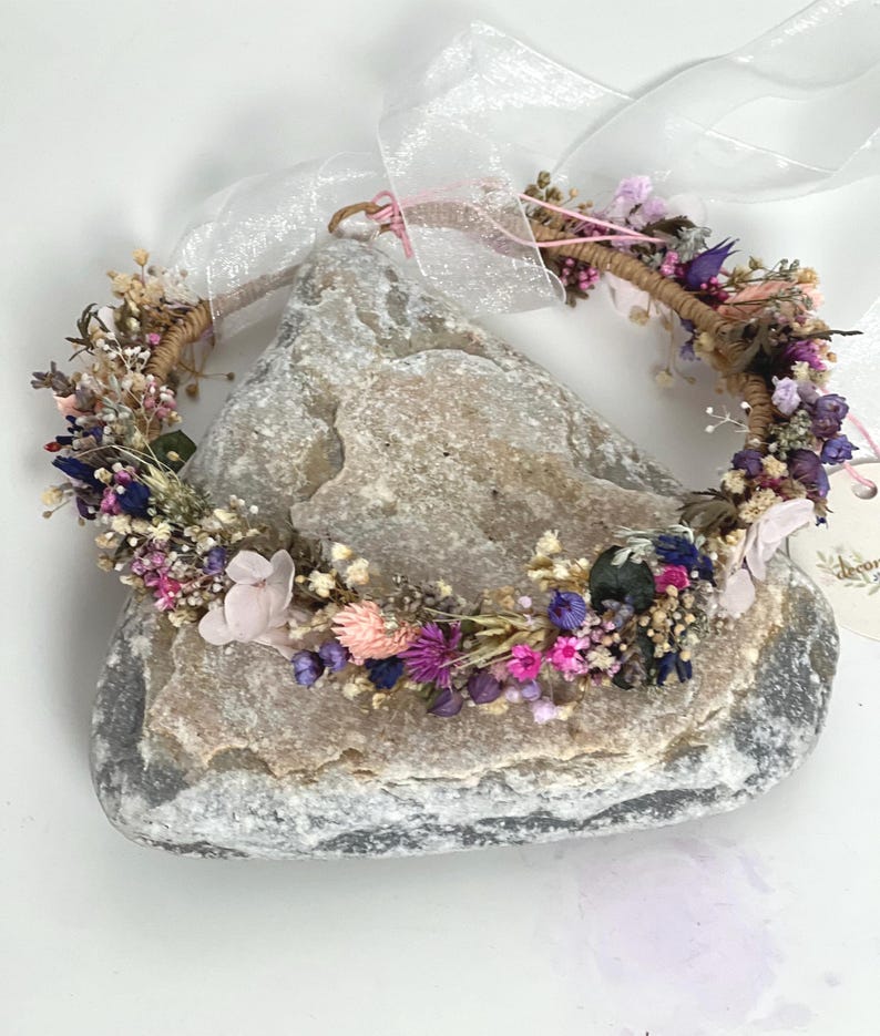 May include: A delicate floral crown made with dried flowers in shades of pink, purple, and white. The crown is wrapped in brown twine and has a light pink ribbon attached.