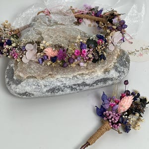 May include: A dried flower crown and matching boutonniere in shades of pink, purple, and white. The crown is made with a natural twine base and the boutonniere is attached to a brown twine stem.