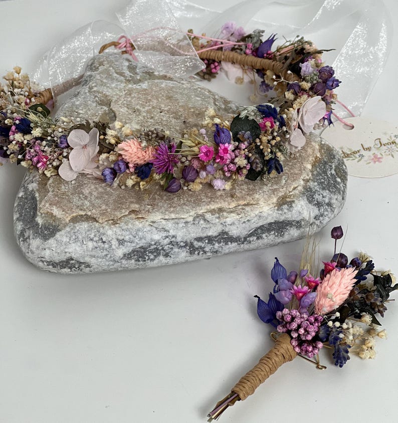 May include: A dried flower crown and matching boutonniere in shades of pink, purple, and white. The crown is made with a natural twine base and the boutonniere is attached to a brown twine stem.