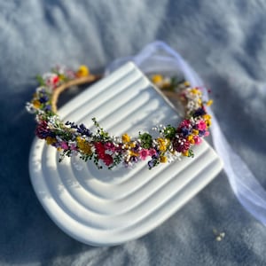 May include: A flower crown made with dried flowers in shades of pink, yellow, and purple. The crown is on a white ceramic base.
