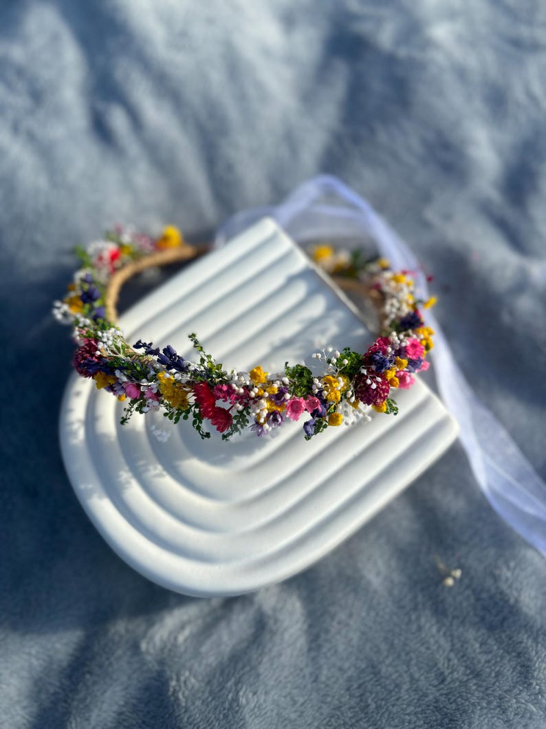 May include: A flower crown made with dried flowers in shades of pink, yellow, and purple. The crown is on a white ceramic base.