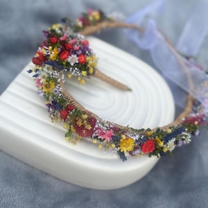 May include: A flower crown made with dried flowers in shades of red, yellow, blue, and white. The crown is made with a natural twine base and is decorated with a variety of small flowers.