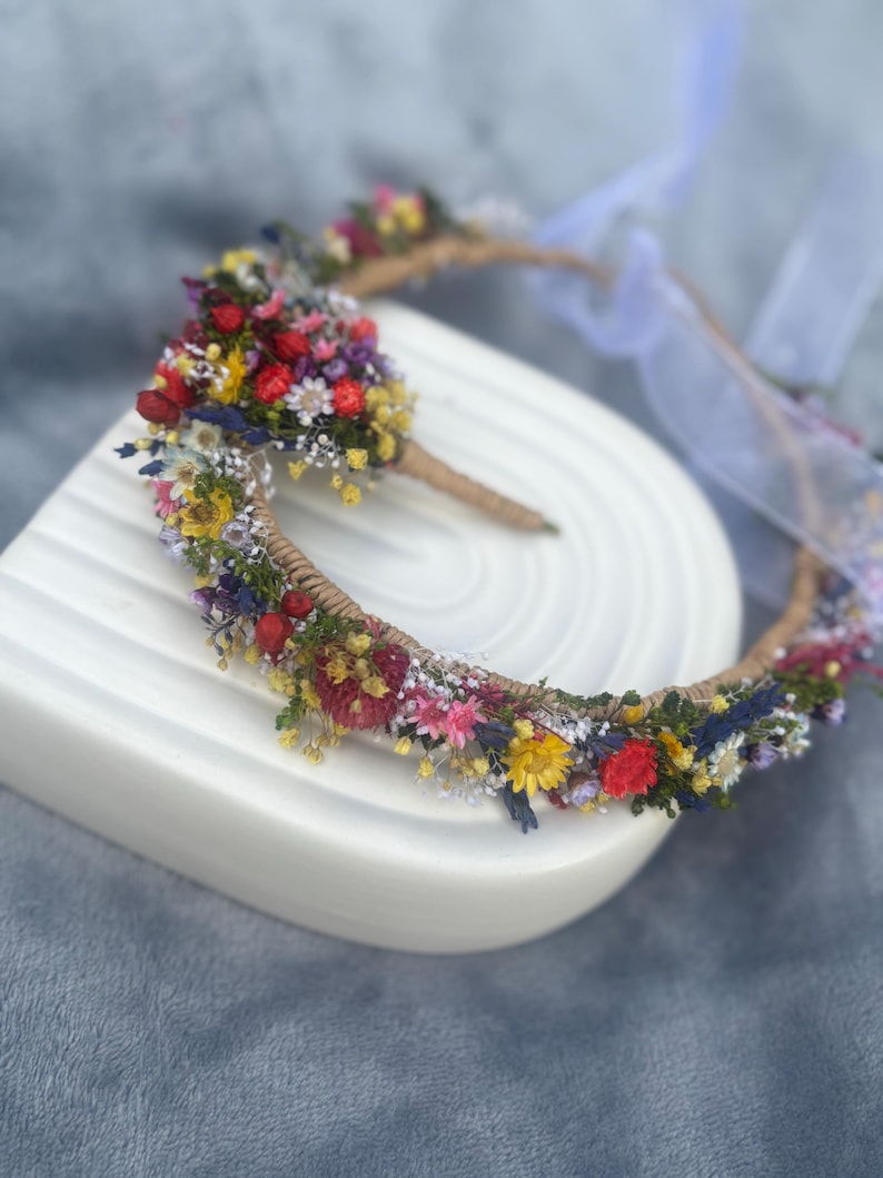 May include: A flower crown made with dried flowers in shades of red, yellow, blue, and white. The crown is made with a natural twine base and is decorated with a variety of small flowers.