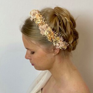 Pode incluir: Uma coroa de flores feita de flores secas em tons de p&ecirc;ssego, creme e bege. A coroa &eacute; usada por uma pessoa com cabelo loiro.
