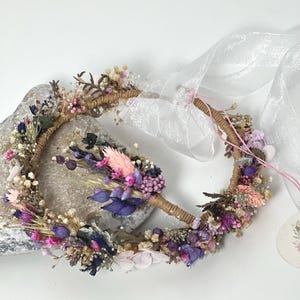 May include: A delicate floral crown made with dried flowers in shades of pink, purple, and white. The crown is made on a brown wire base and is adorned with a sheer white ribbon.