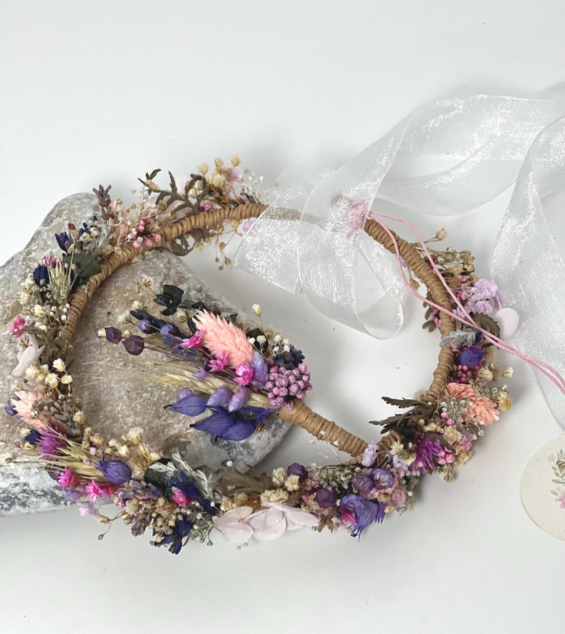 May include: A delicate floral crown made with dried flowers in shades of pink, purple, and white. The crown is made on a brown wire base and is adorned with a sheer white ribbon.