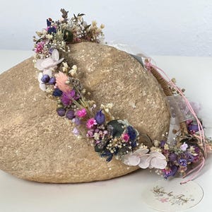 May include: A delicate flower crown with dried flowers in shades of pink, purple, and white. The crown is made with a natural, woven base and is adorned with a variety of small flowers and greenery.