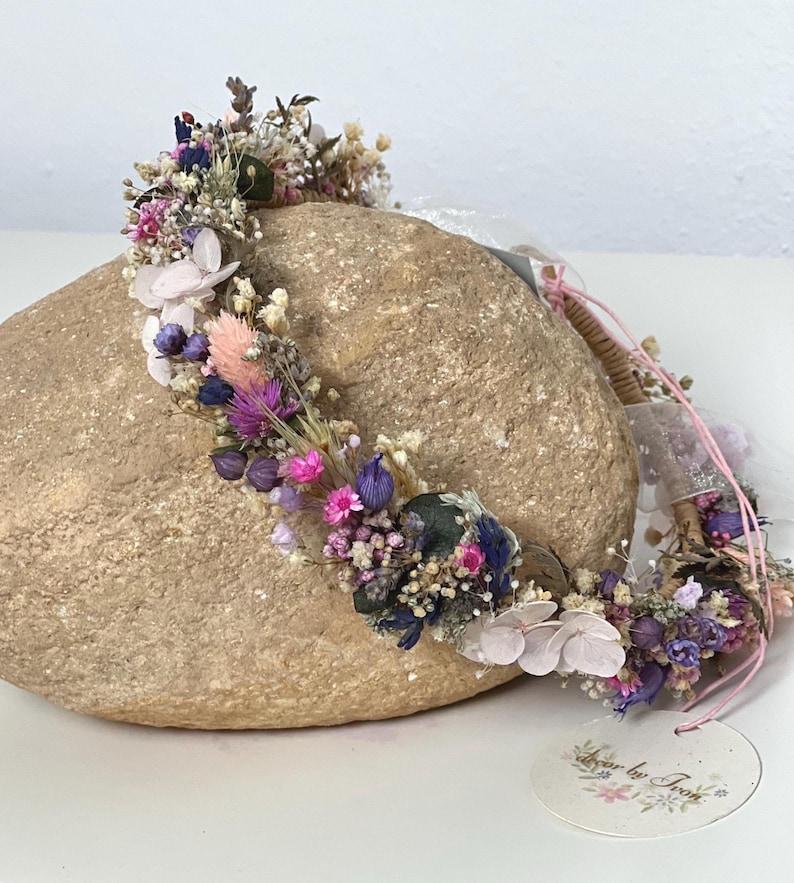May include: A delicate flower crown with dried flowers in shades of pink, purple, and white. The crown is made with a natural, woven base and is adorned with a variety of small flowers and greenery.