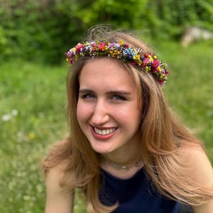 May include: A woman with long blonde hair wearing a flower crown made of small colorful flowers. The crown is made of a variety of flowers, including pink, purple, yellow, and blue.