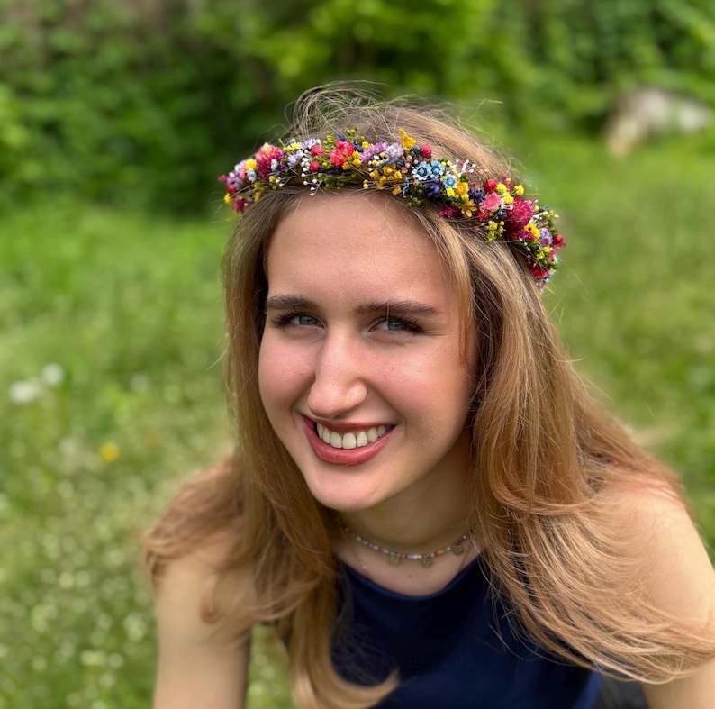May include: A woman with long blonde hair wearing a flower crown made of small colorful flowers. The crown is made of a variety of flowers, including pink, purple, yellow, and blue.