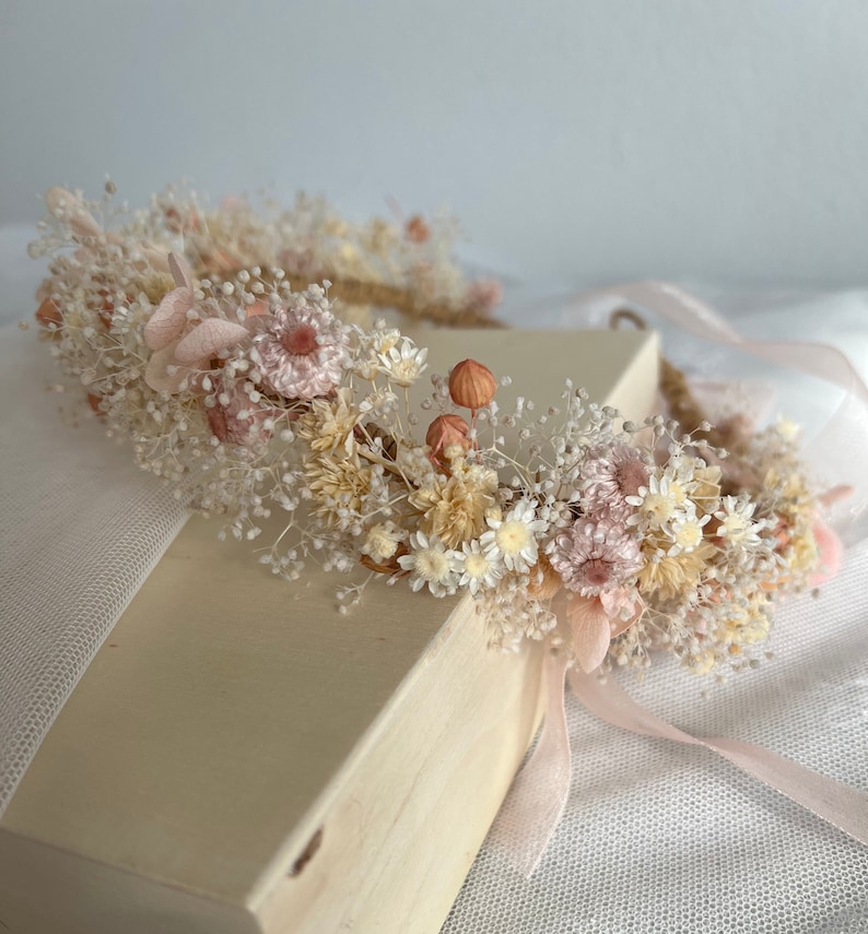 Pode incluir: Uma coroa floral delicada feita de flores secas em tons de rosa e creme. A coroa &eacute; adornada com pequenas flores brancas e rosas, bem como folhas e caules secos.