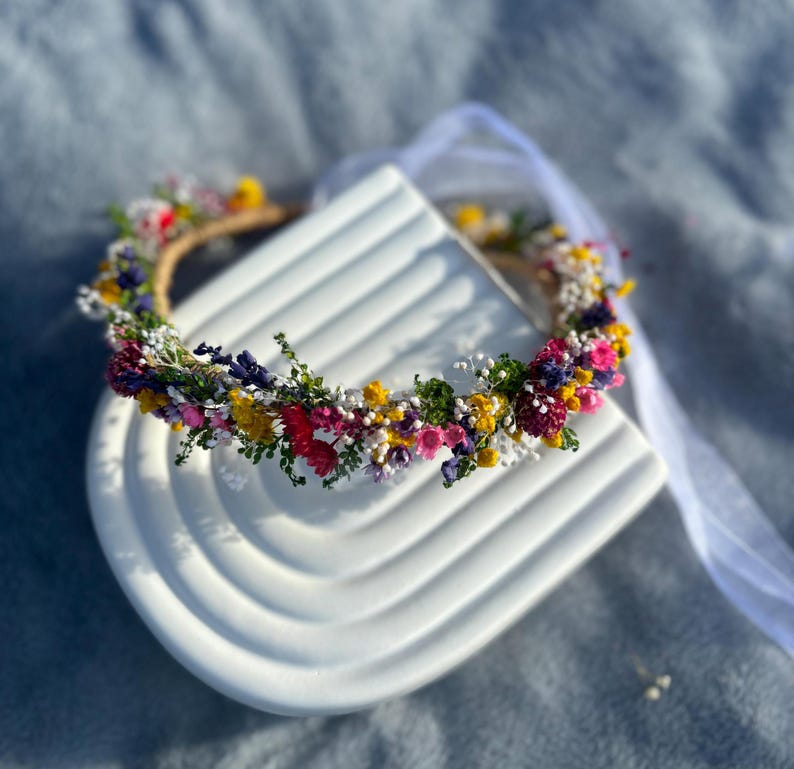 May include: A flower crown made of dried flowers in shades of pink, yellow, white, and purple. The crown is on a white surface.