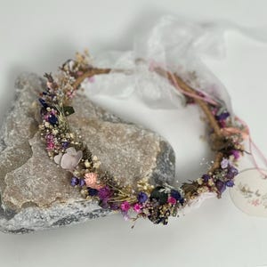 May include: A delicate floral crown made with dried flowers in shades of pink, purple, and white. The crown is made on a natural twig base and is adorned with small, delicate flowers.