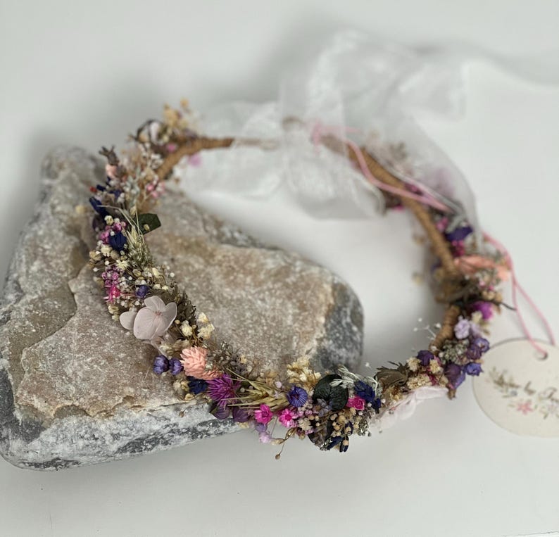 May include: A delicate floral crown made with dried flowers in shades of pink, purple, and white. The crown is made on a natural twig base and is adorned with small, delicate flowers.