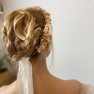 Pode incluir: Uma mulher com cabelo loiro usando uma coroa de flores com uma fita branca amarrada na parte de tr&aacute;s da cabe&ccedil;a. A coroa &eacute; feita de flores secas em tons de bege e rosa.