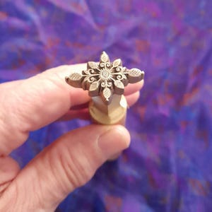 May include: A detailed metal stamp with a floral design. The stamp features a central flower with surrounding petal-like shapes. The stamp is held against a purple fabric background.