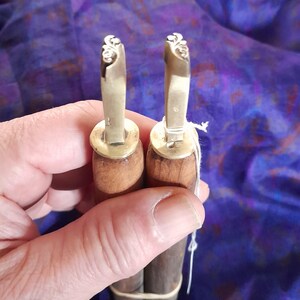 May include: Two leather working tools with wooden handles and brass heads. The brass heads feature a decorative floral design. The handles are a dark brown colour. The tools are held together with a rubber band and white string.