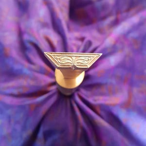 May include: A gold-coloured stamp with a triangular head featuring an ornate, symmetrical floral design. The stamp is set against a blurred purple fabric background. The stamp's handle is cylindrical. The design creates a contrast in textures and colours.