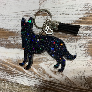 May include: A German Shepherd dog keychain with a black glitter finish. The keychain includes a silver keyring, a paw print charm, and a black tassel. The dog silhouette is detailed and stands out against the wooden background.