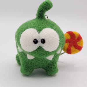 Pode incluir: Um monstro verde de feltro feito à mão com olhos brancos grandes, dentes brancos pequenos e uma cauda enrolada. Ele segura um pirulito colorido com listras vermelhas e amarelas. O brinquedo é um personagem fantasioso.