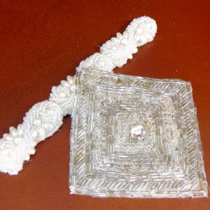 以下が含まれることがあります： 白いビーズのハンドバッグ、持ち手付き。 バッグは正方形で、中央に宝石があり、ビーズの持ち手が付いています。 バッグは小さな銀色のビーズと白いビーズで作られています。 持ち手には花のディテールがあります。