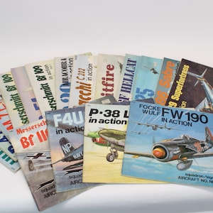 Könnte beinhalten: Eine Sammlung von Vintage-Luftfahrtbüchern, auf weißem Hintergrund aufgefächert. Die Cover zeigen Illustrationen verschiedener Flugzeuge, darunter die Focke Wulf FW 190, P-38 L und andere. Titel sind "In Action" und Flugzeugnamen.