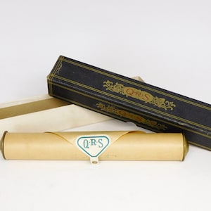 May include: A vintage black rectangular box with gold detailing and the letters "QRS". A cream-coloured box and a rolled parchment with a "QRS" label are also present. The items are on a white background.