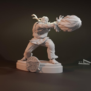 Può includere: Una statuetta bianca stampata in 3D di un personaggio di Street Fighter, Ryu, in una posa di combattimento con un effetto di palla di fuoco. La statuetta è in piedi su una base con il logo di Street Fighter.