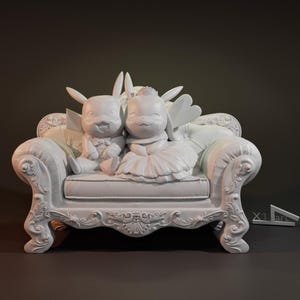 Könnte beinhalten: Weiße Keramikfigur eines Paares von hasenartigen Kreaturen, die auf einem weißen, verzierten Sofa sitzen. Die weibliche Figur trägt ein Hochzeitskleid und die männliche Figur trägt einen Smoking.