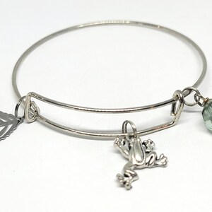 Könnte beinhalten: Ein silberfarbenes, erweiterbares Armband mit drei hängenden Anhängern: einem flachen Metallblatt, einer kleinen dreidimensionalen Froschfigur und einer einzelnen, durchscheinenden, hellgrünen, facettierten Glasperle an einem kleinen Metallring.